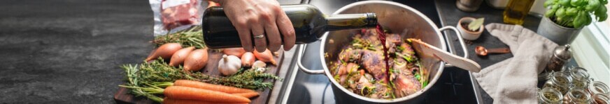 Koken met wijn: een fles rode wijn wordt in een pan geschonken waar al vlees en kruiden gebakken worden. verder liggen er wortels, uien en kruiden op het aanrecht